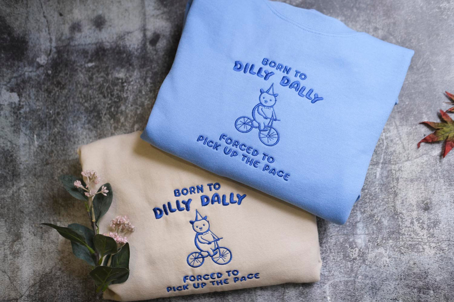 two sweatshirts with a blue and a beige color scheme, each with a unique design and text. The blue sweatshirt has a cartoon image of a cat riding a bicycle and the text "Born to Dilly Dally" and "Forced to Pick Up the Pace". The beige sweatshirt has a similar cartoon image of a cat riding a bicycle and the text "Born to Dilly Dally" and "Forced to Pick Up the Pace".