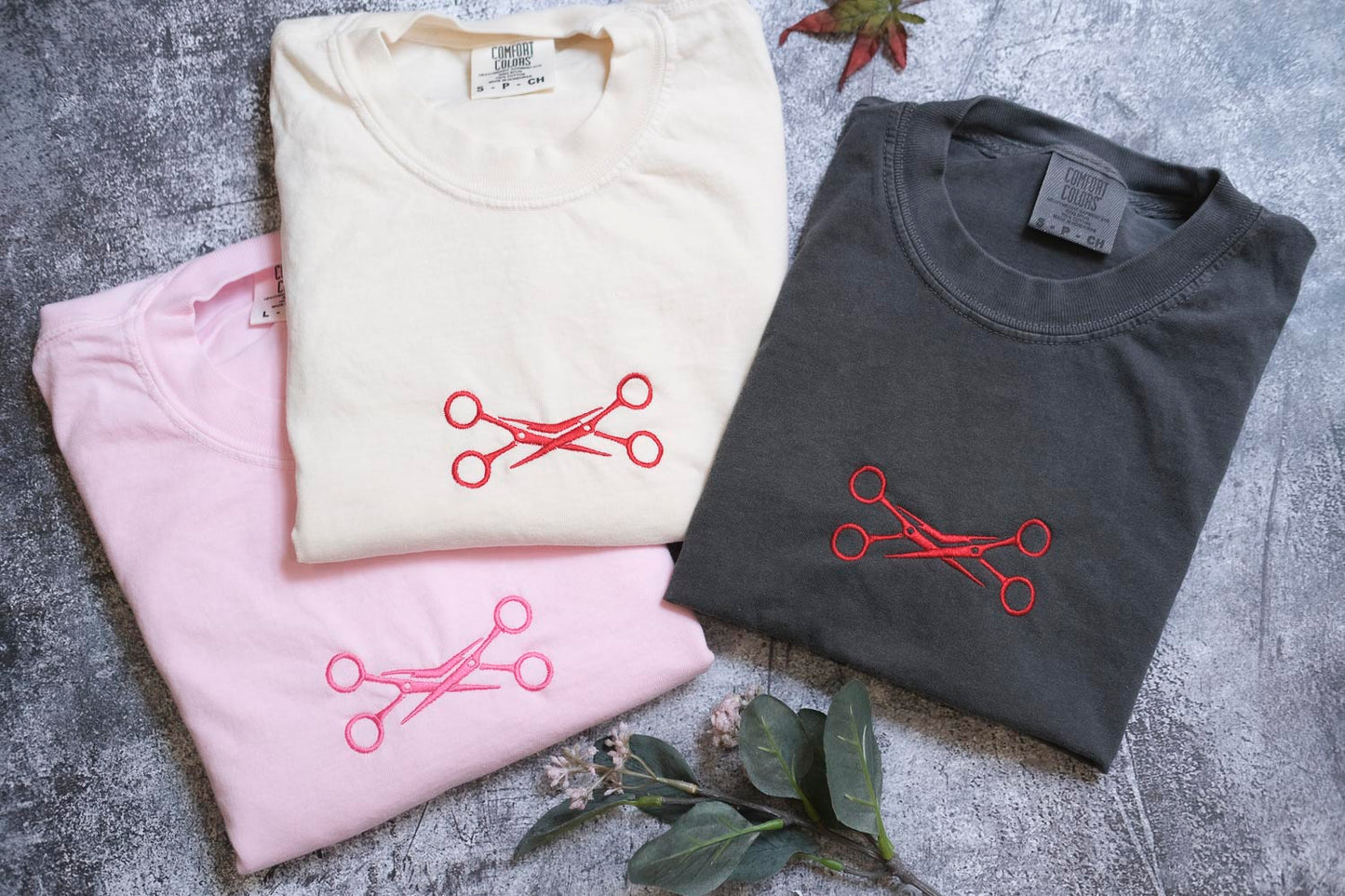 three t-shirts with scissors embroidered on them, arranged on a gray surface.