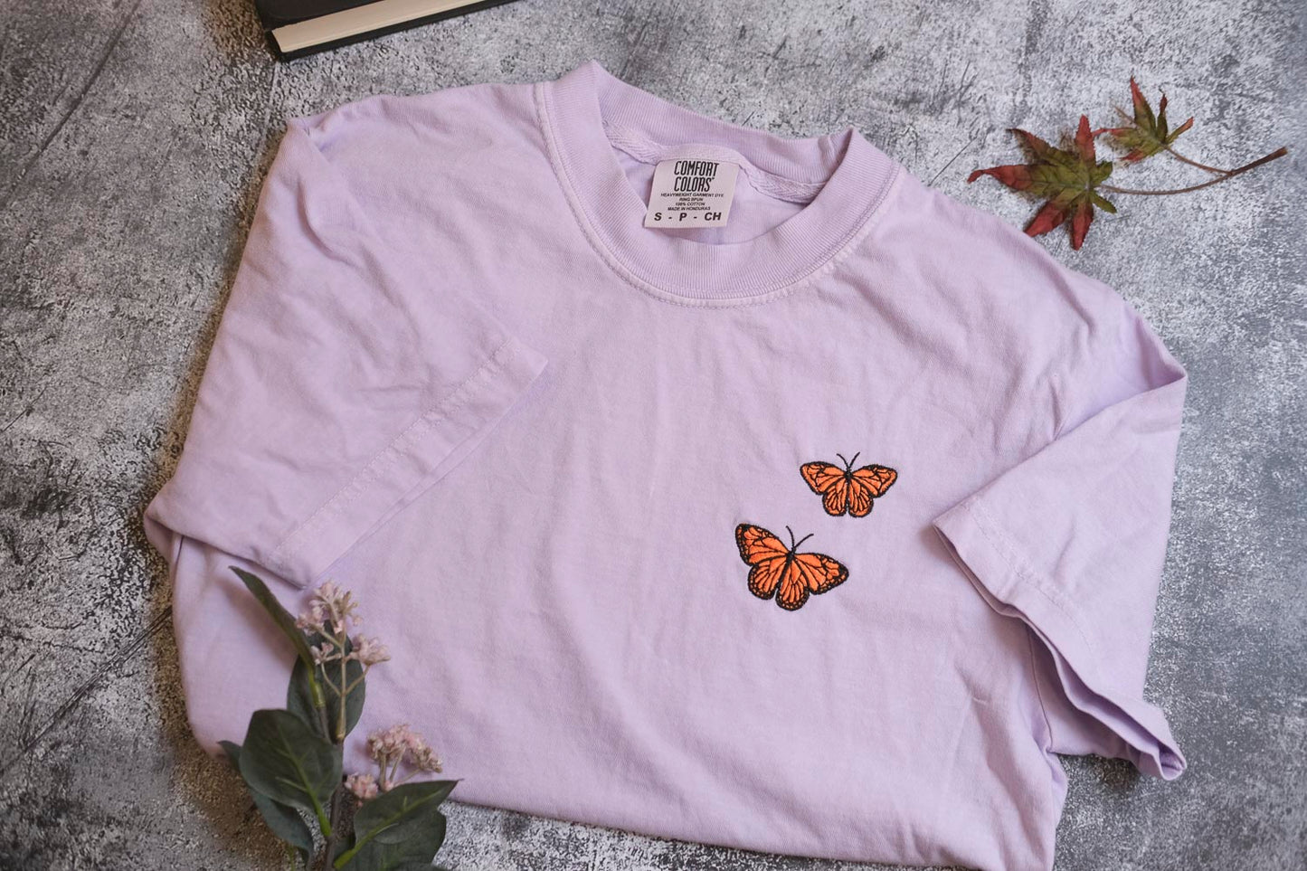 a light purple t-shirt with two orange butterflies printed on it, placed on a gray surface alongside some leaves and a small plant.