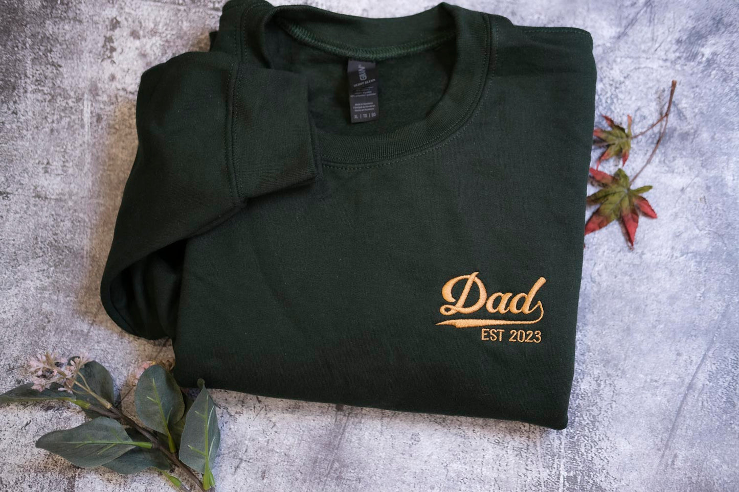 A dark green sweatshirt with the text "DAD EST 2023" printed on it, along with a small gold-colored logo.
