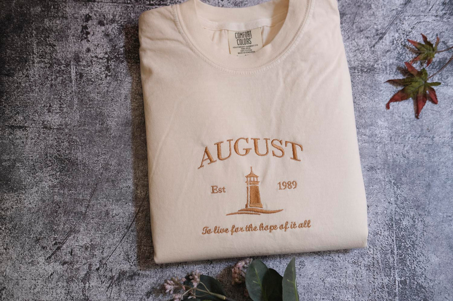 a white t-shirt with a printed design on it, placed on a textured gray surface. The design features a lighthouse and the text "AUGUST Est. 1989" along with the phrase "To live for the hope of it all".