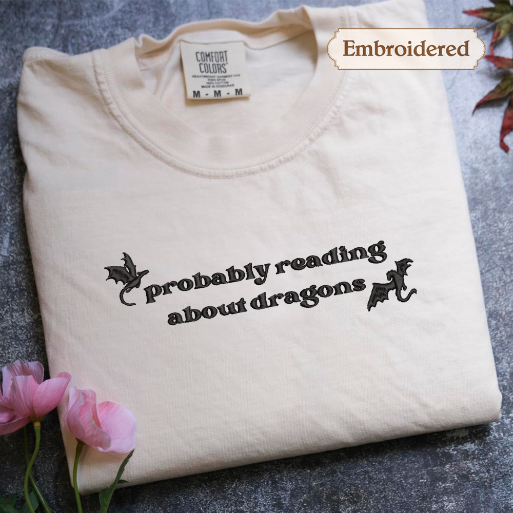 A white t-shirt with the text "probably reading about dragons" embroidered on it, along with a dragon design. The t-shirt is placed on a gray surface, and there are pink flowers nearby.