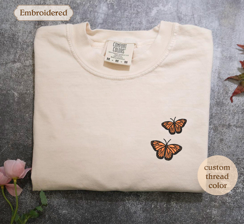 a white t-shirt with a small embroidered design of two orange butterflies on the left side of the chest area.