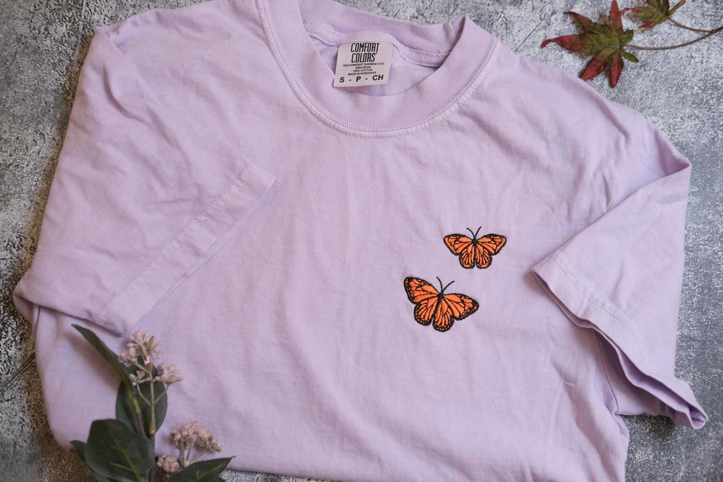 A light purple t-shirt with two orange butterflies printed on it, placed on a gray surface with some greenery and a small plant.