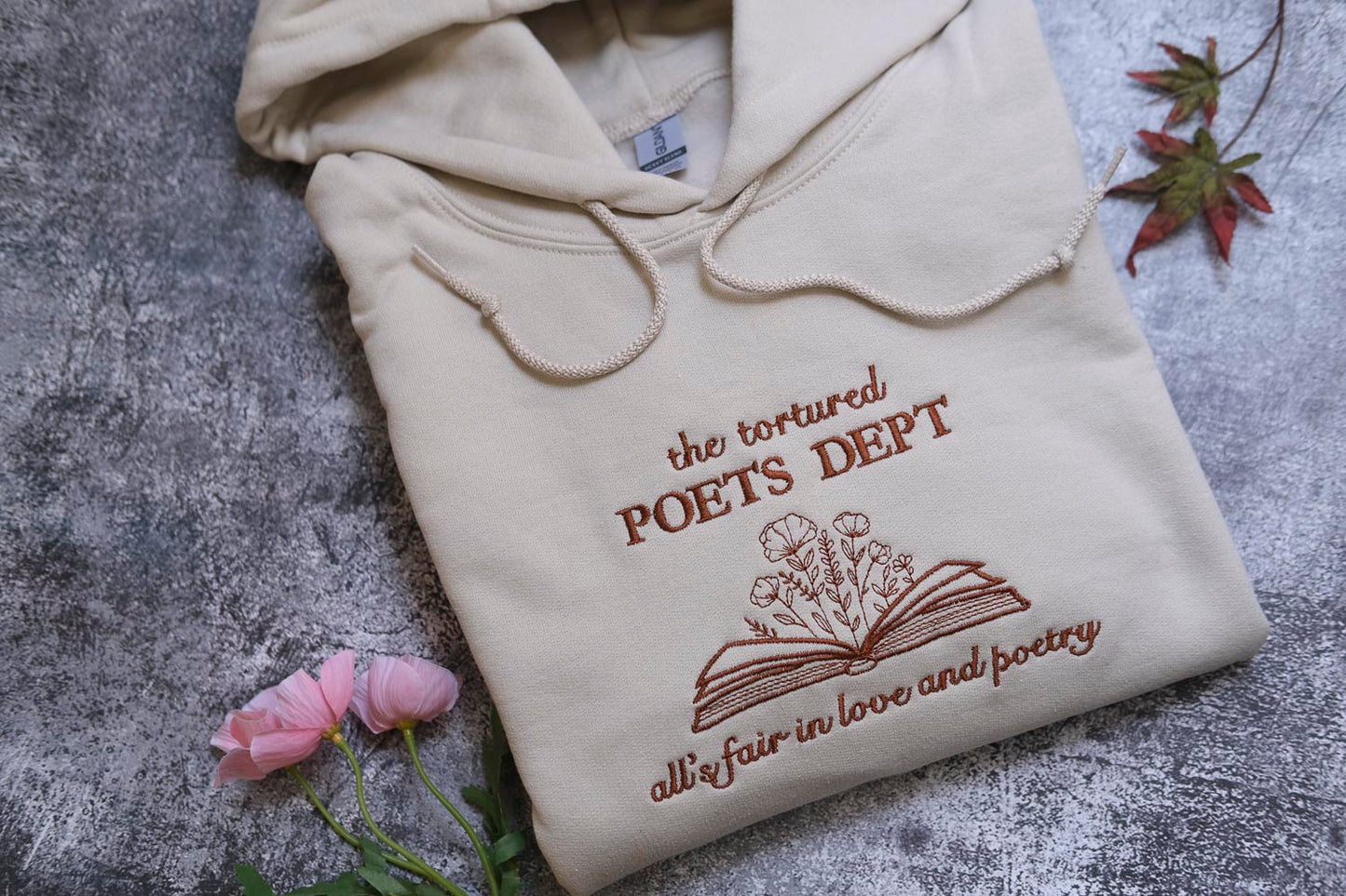 a beige hoodie with the text "the tortured POET'S DEPT. all fair in love and poetry" printed on it, along with an illustration of an open book with flowers. The hoodie is placed on a gray surface, and there are some pink flowers and a leaf nearby.