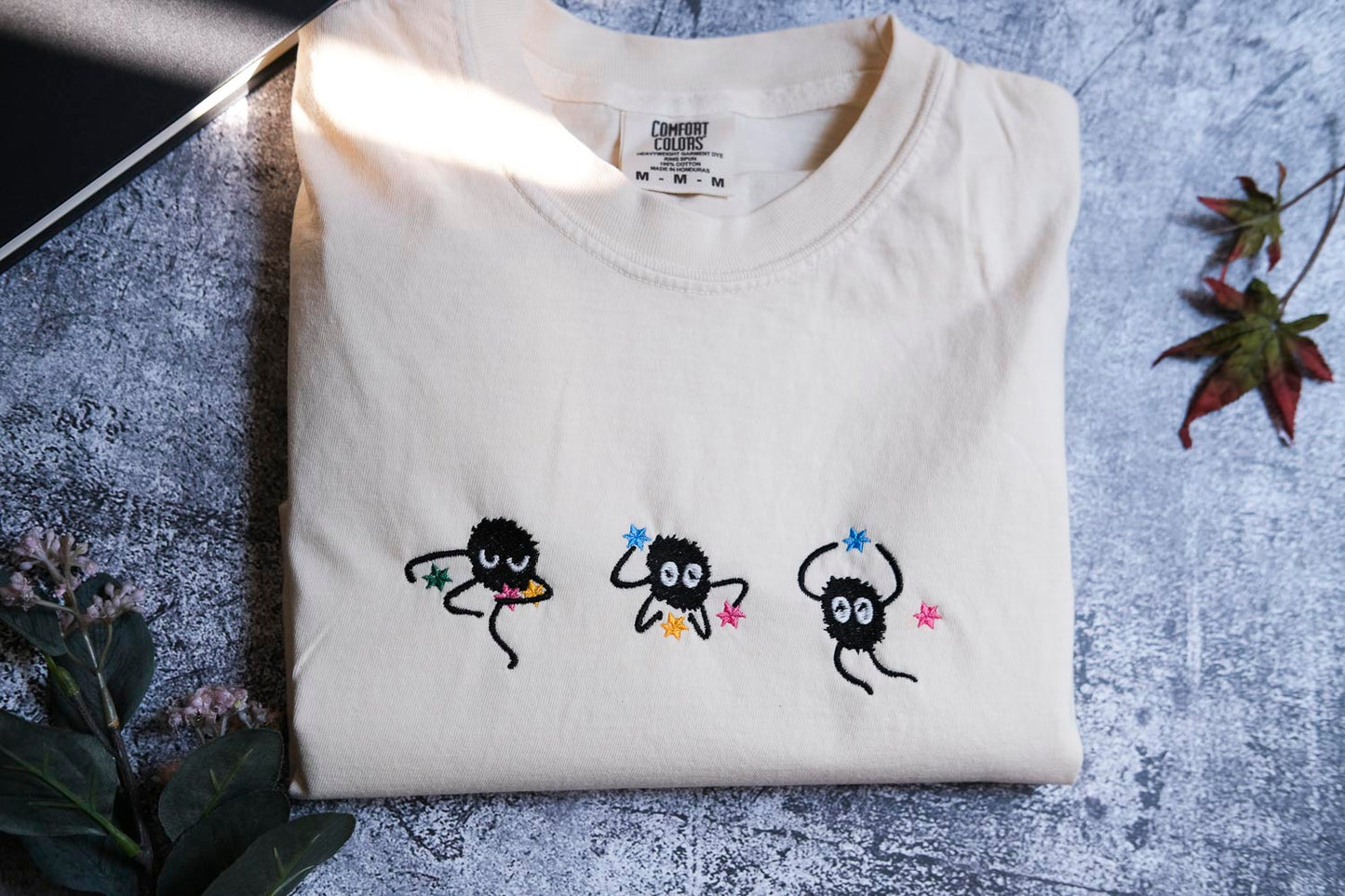 a white t-shirt with three small black creatures with colorful eyes and arms, arranged in a row on the front of the shirt.