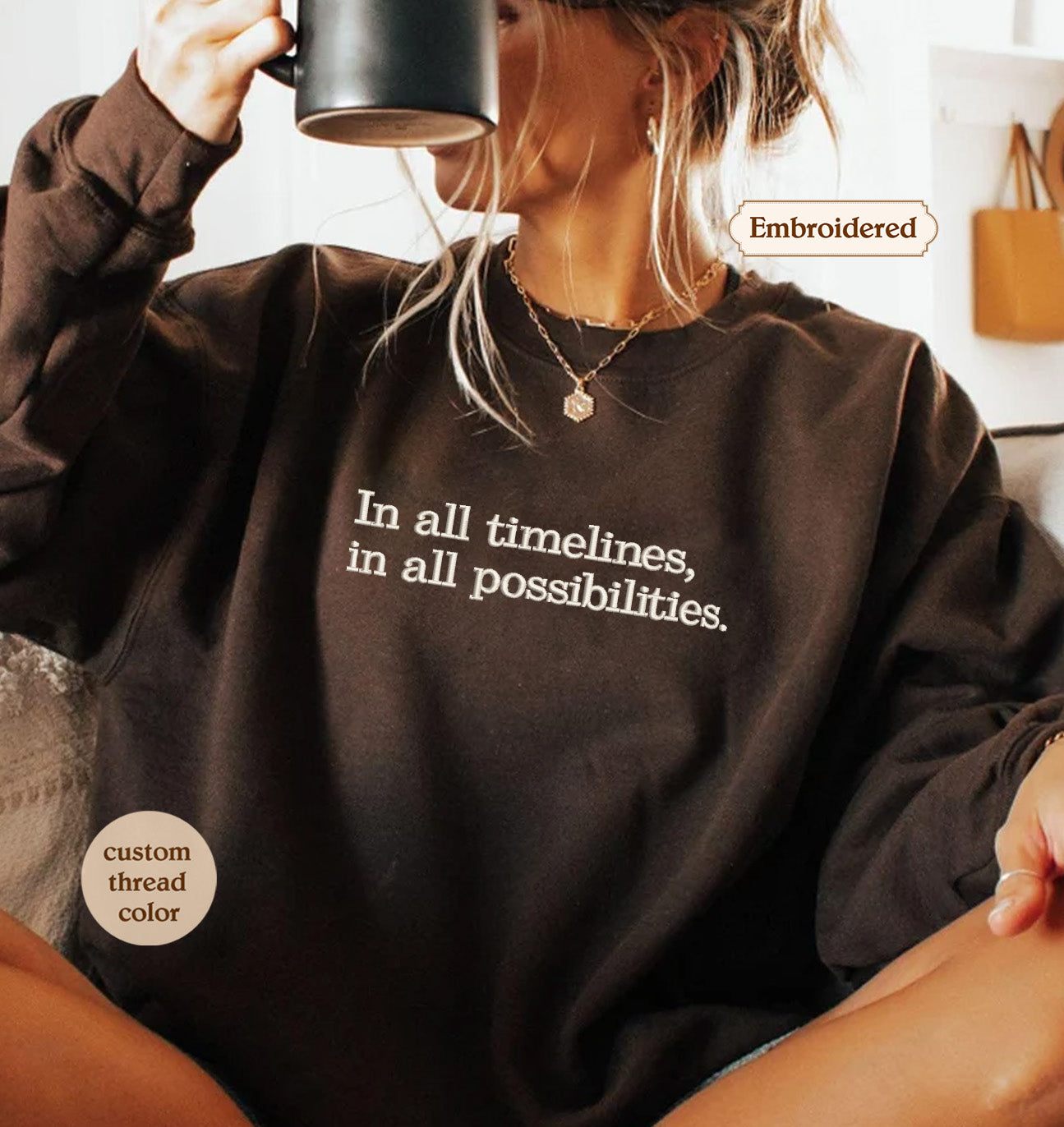 A woman wearing a brown sweatshirt with the text "In all timelines, in all possibilities" printed on it, holding a coffee mug and sitting on a couch.