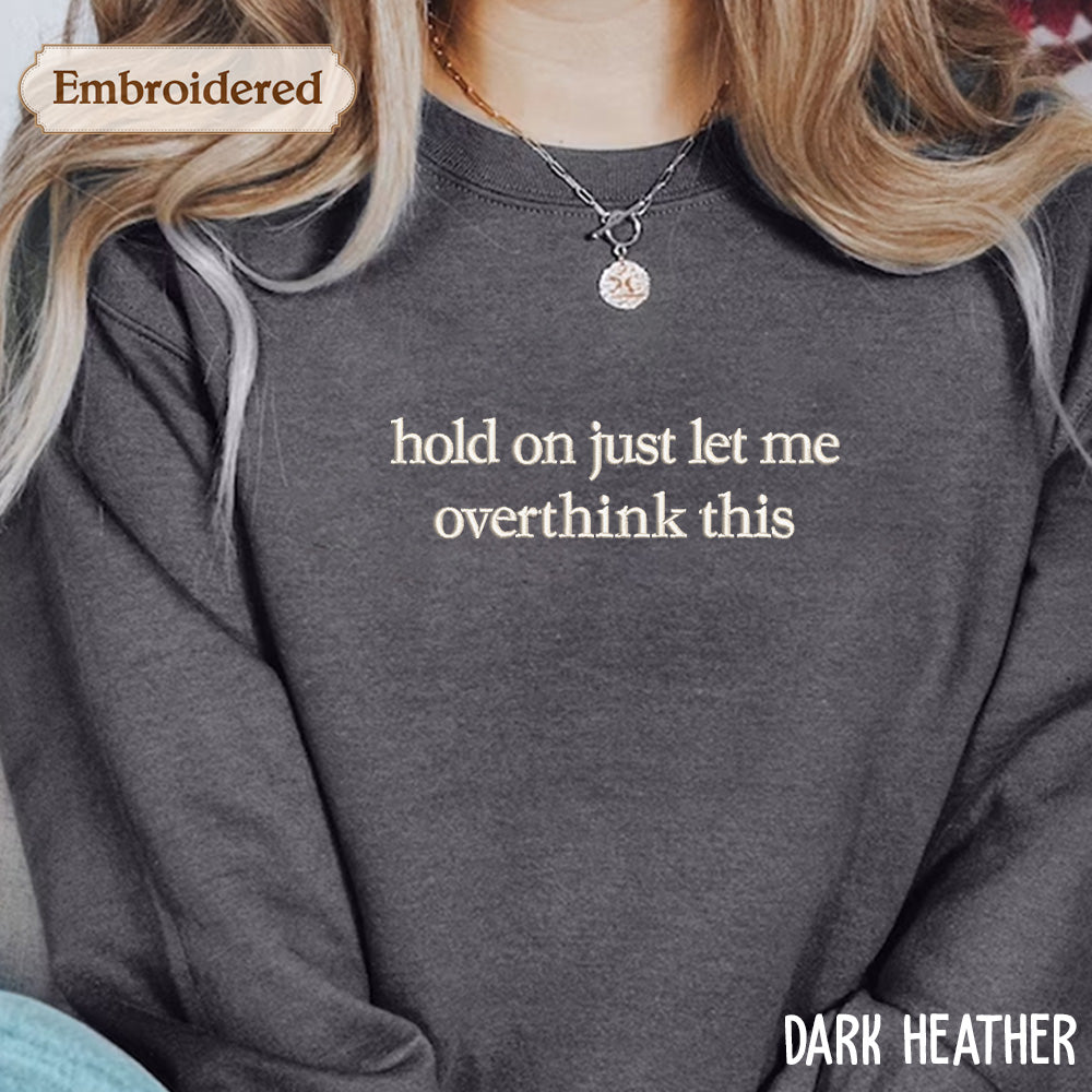 A person wearing a gray sweatshirt with the text "hold on just let me overthink this" embroidered on it.