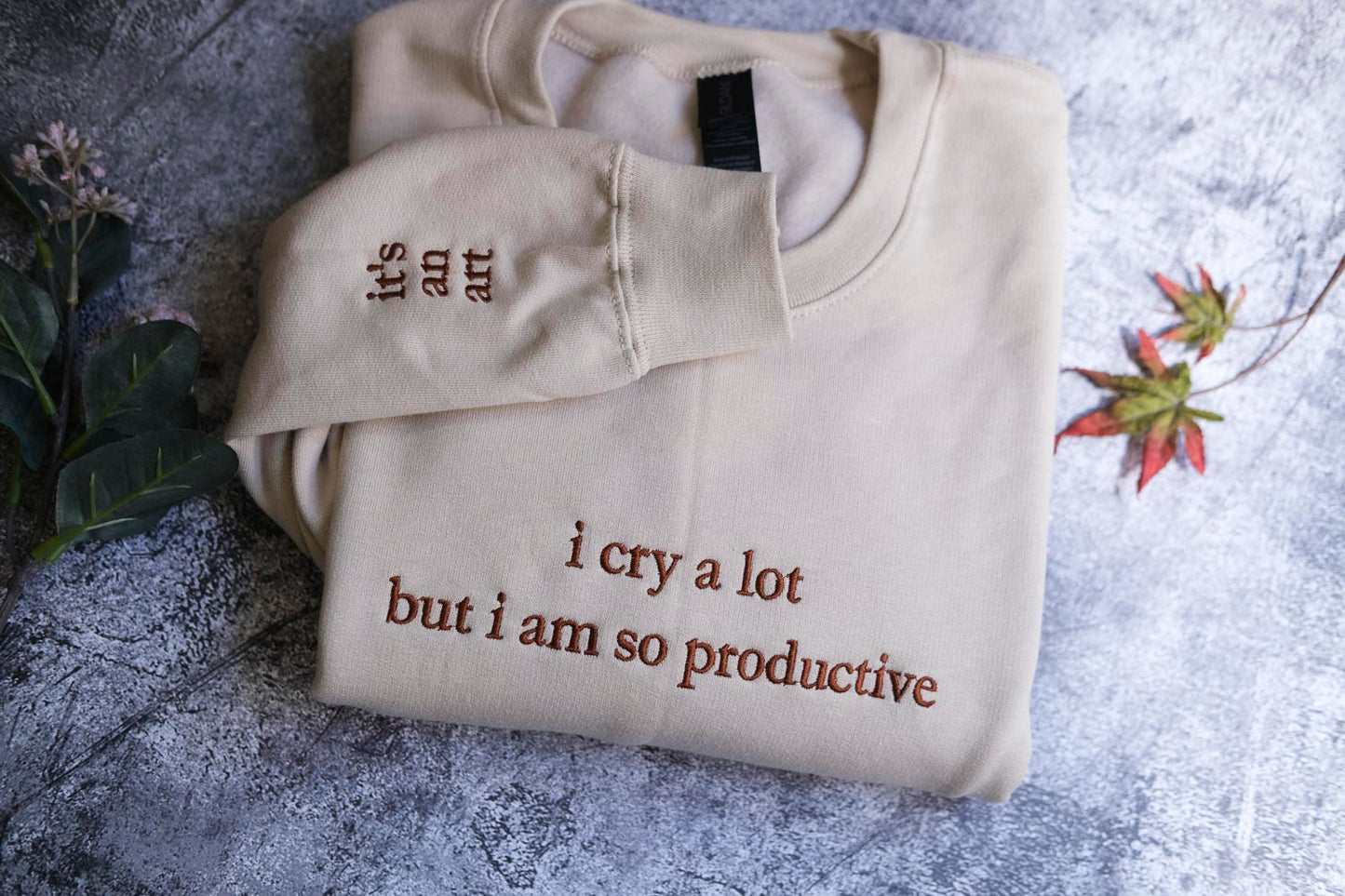 a beige sweater with the text "It's an art" and "I cry a lot but I am so productive" printed on it, along with a plant and a leaf in the background.