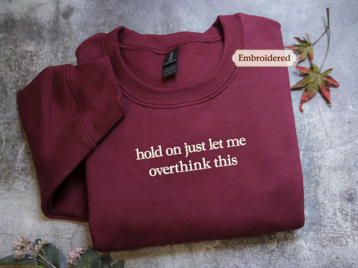 A burgundy sweatshirt with the text "hold on just let me overthink this" embroidered on it, along with a small embroidered label that says "Embroidered".