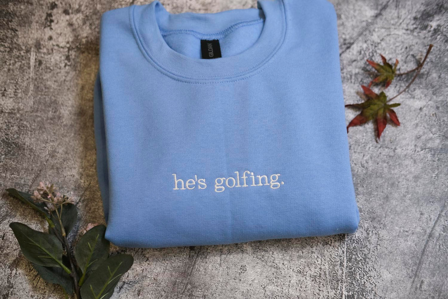 A blue sweatshirt with the text "he's golfing" printed on it, along with some leaves and a small plant.