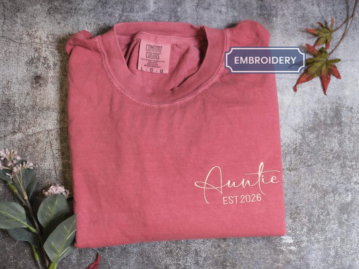 a pink t-shirt with the text "Auntie EST 2026" printed on it, along with some leaves and flowers.
