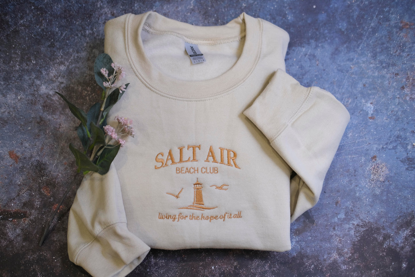 A beige sweater with the text "SALT AIR BEACH CLUB" and a lighthouse graphic printed on it, along with the phrase "Living for the hope of all" printed below.