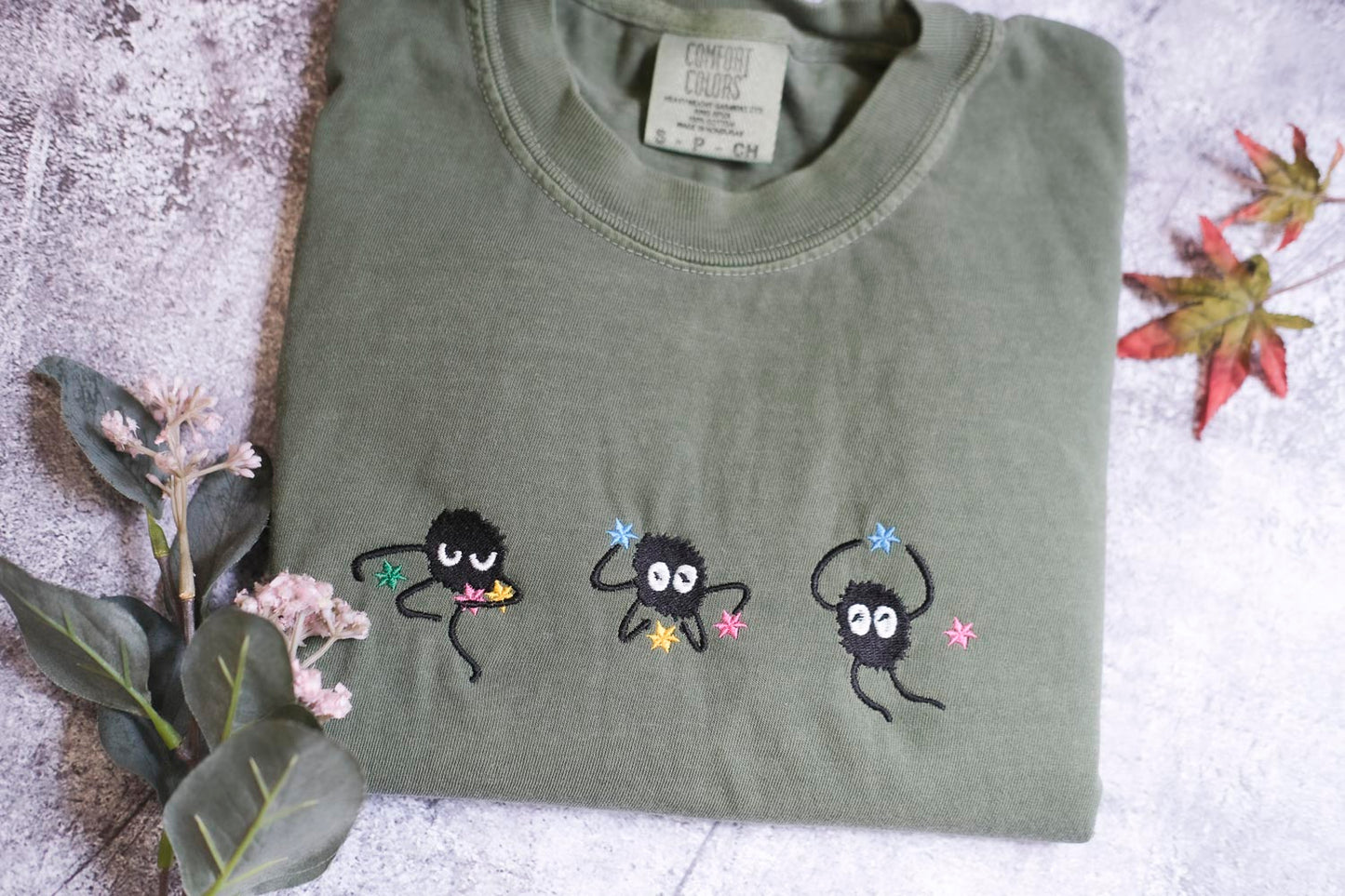 a green t-shirt with three small black creatures with white eyes and colorful accents, such as flowers, on it. The t-shirt is placed on a surface with some leaves and flowers, creating a natural and whimsical setting.
