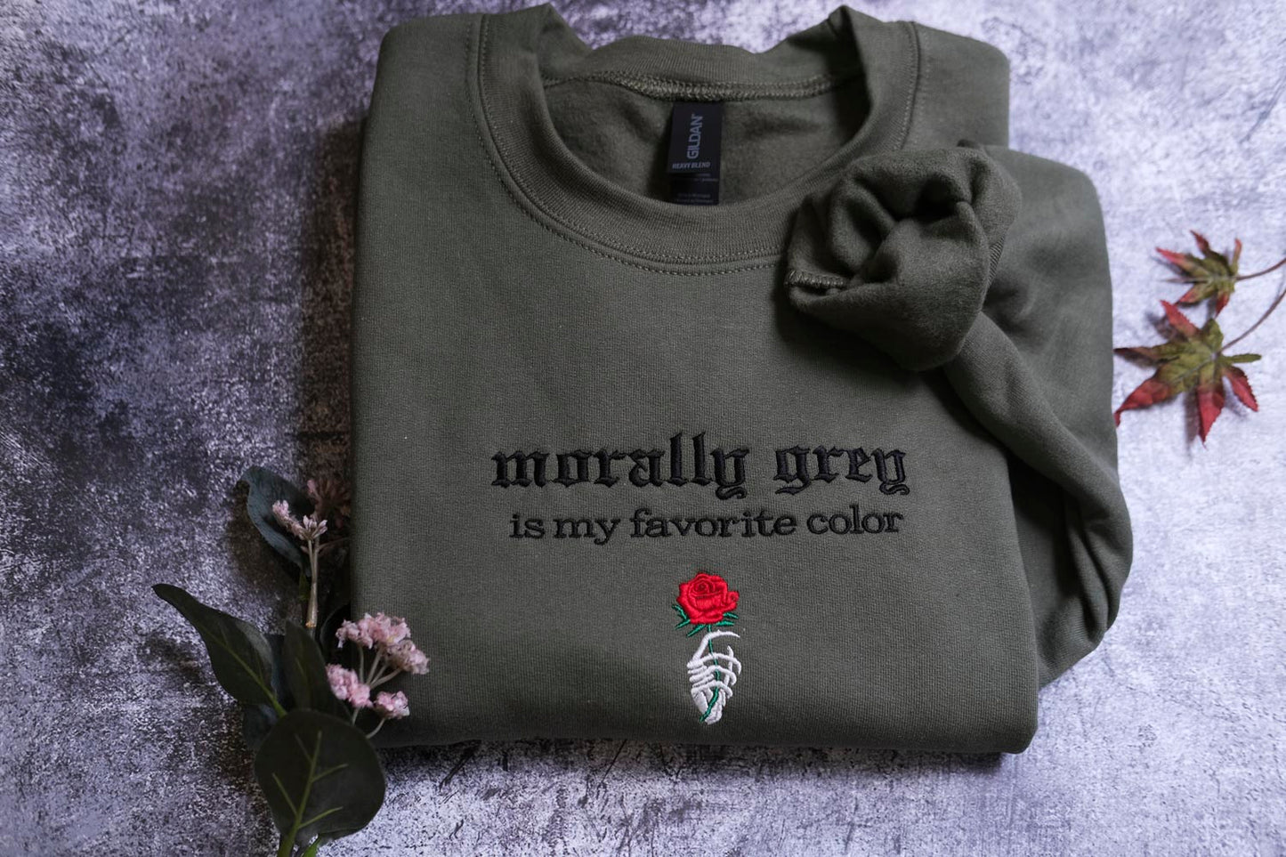 a dark green t-shirt with a black text design and a red rose embroidered on it, placed on a gray surface.