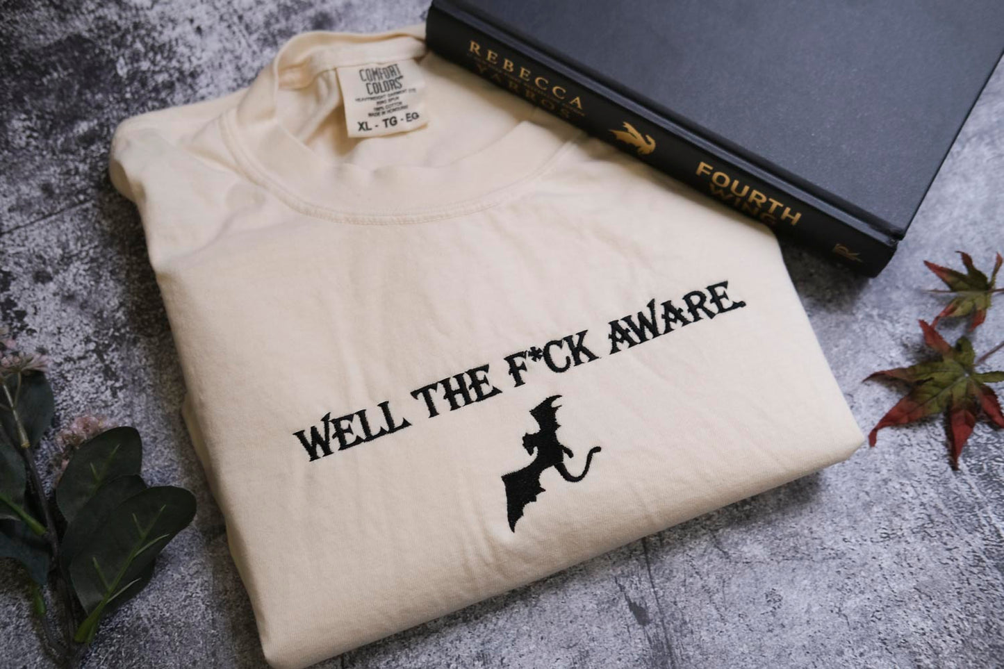 a white t-shirt with the text "WELL THE F*CK AWARE" printed on it, along with a graphic of a bat. The t-shirt is placed next to a black book titled "FOURTH" and some leaves.