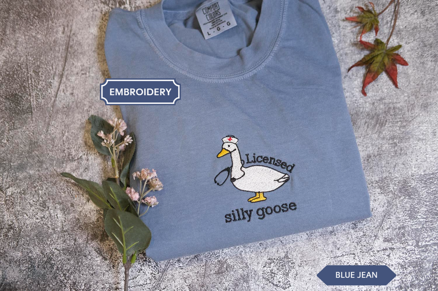 A blue t-shirt with a cartoon image of a white goose wearing a nurse's cap and the text "Licensed silly goose" printed on it, along with some flowers and a leaf.
