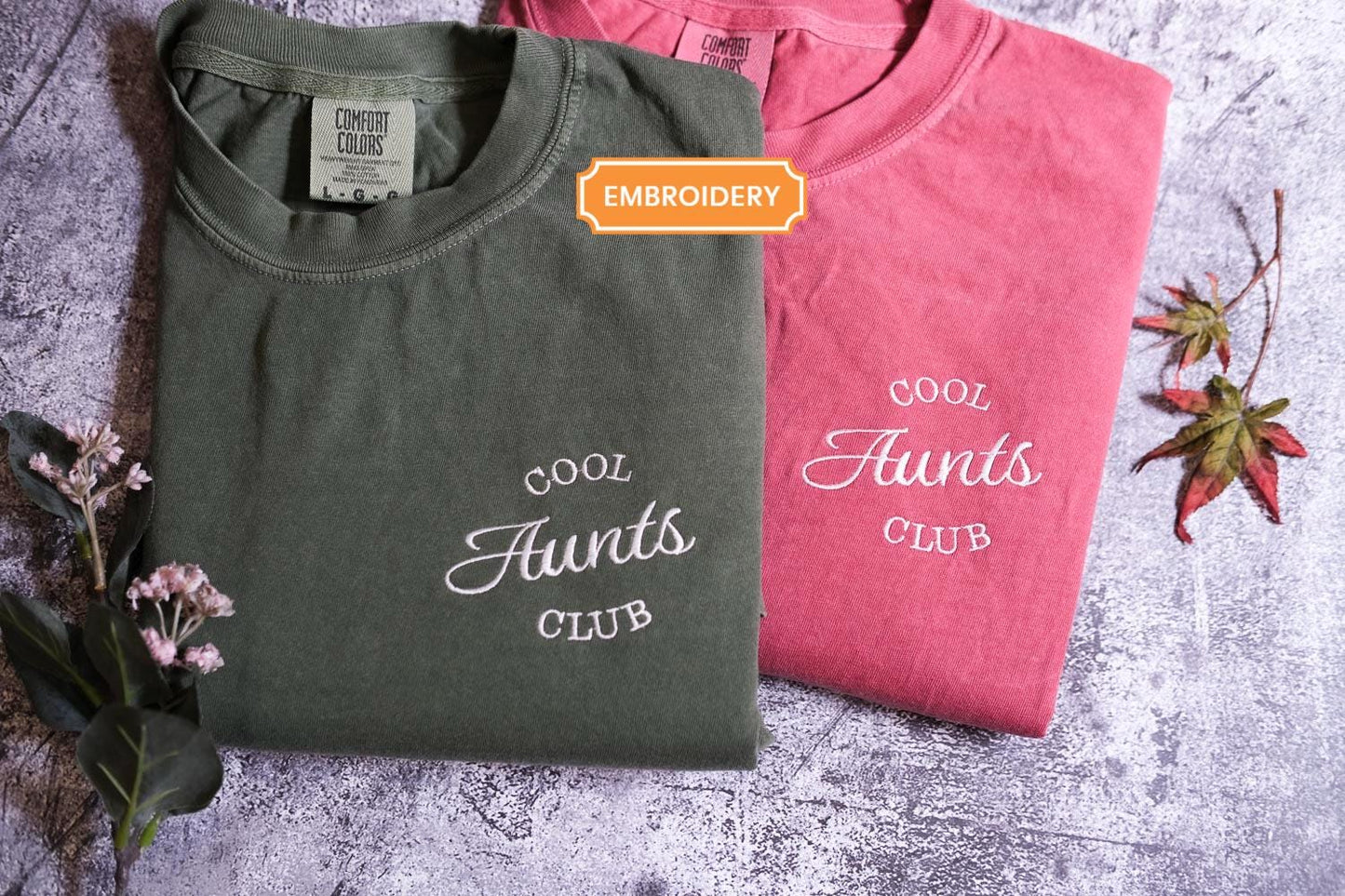 Two t-shirts are displayed on a gray surface, one in a dark green color and the other in a pink color. The dark green t-shirt has the text "Cool Aunt's Club" printed on it, while the pink t-shirt has the text "Cool Aunt's Club" printed on it. There are also some pink flowers and a fallen leaf in the background.