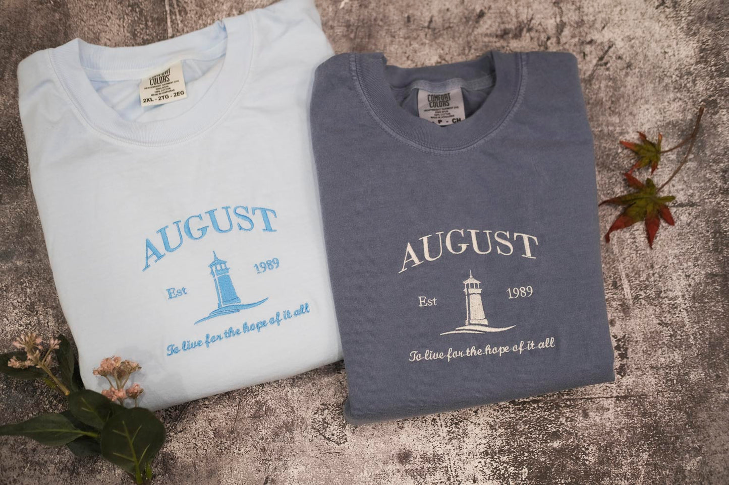 two t-shirts with the same design, laid out on a textured surface. The t-shirts are white and gray, and they feature a lighthouse and the text "August" printed on them.