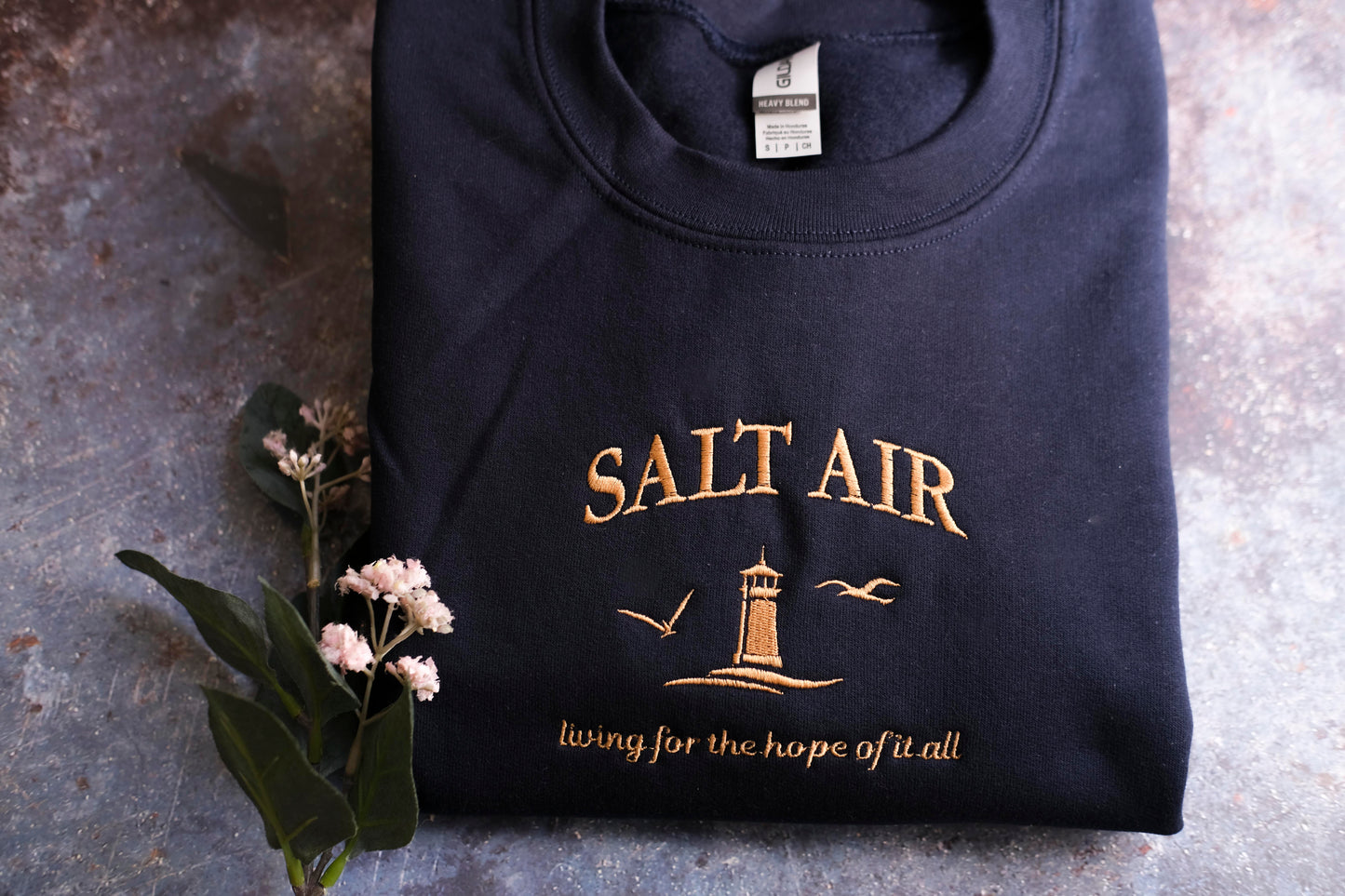 A navy blue sweatshirt with the text "SALT AIR" and a lighthouse graphic printed on it, along with the phrase "living for the hope of all".
