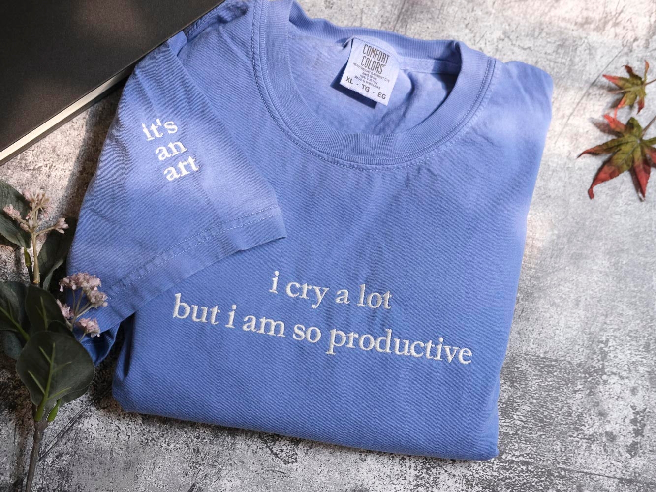A blue t-shirt with white text printed on it, lying on a surface with some leaves and a book.