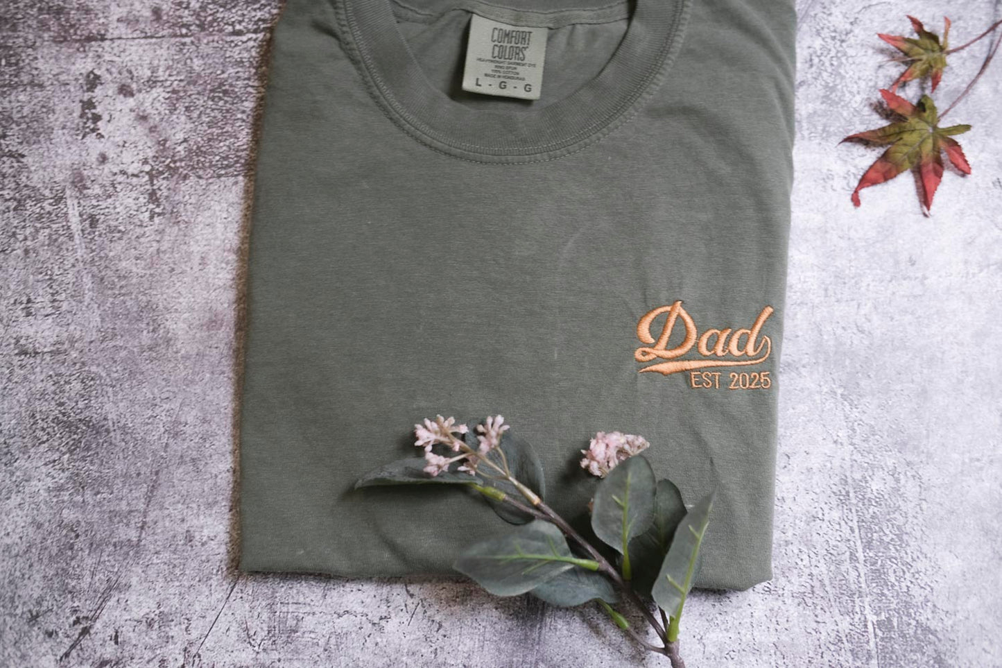 a gray t-shirt with a printed design, a small branch with white flowers, and a leaf on a textured surface.
