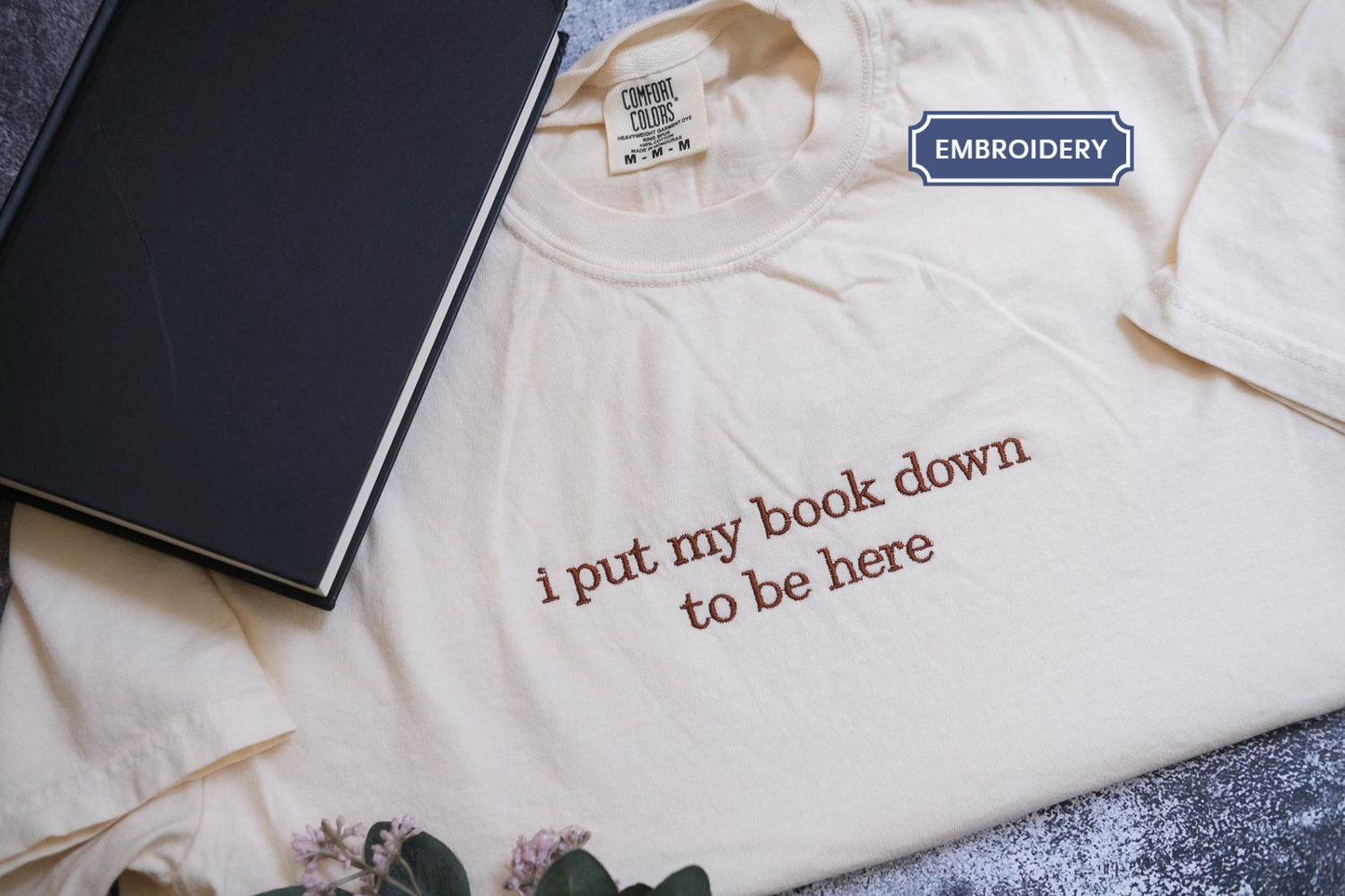 a white t-shirt with the text "I put my book down to be here" printed on it, along with a black book and some flowers.