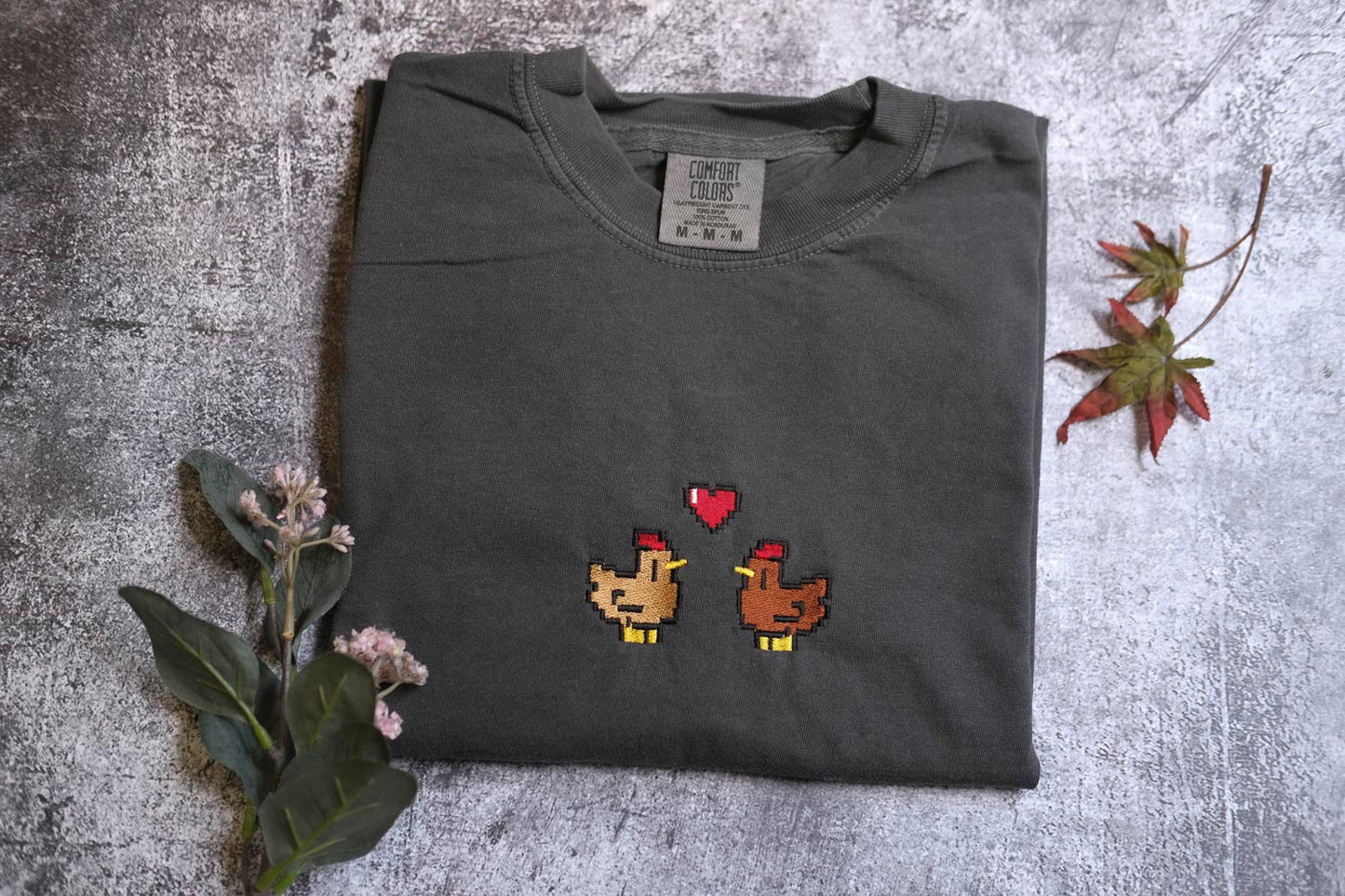 a gray t-shirt with a pixelated design of two chickens, one brown and one yellow, with a heart between them. The t-shirt is placed on a gray surface, and there are some flowers and a leaf nearby.