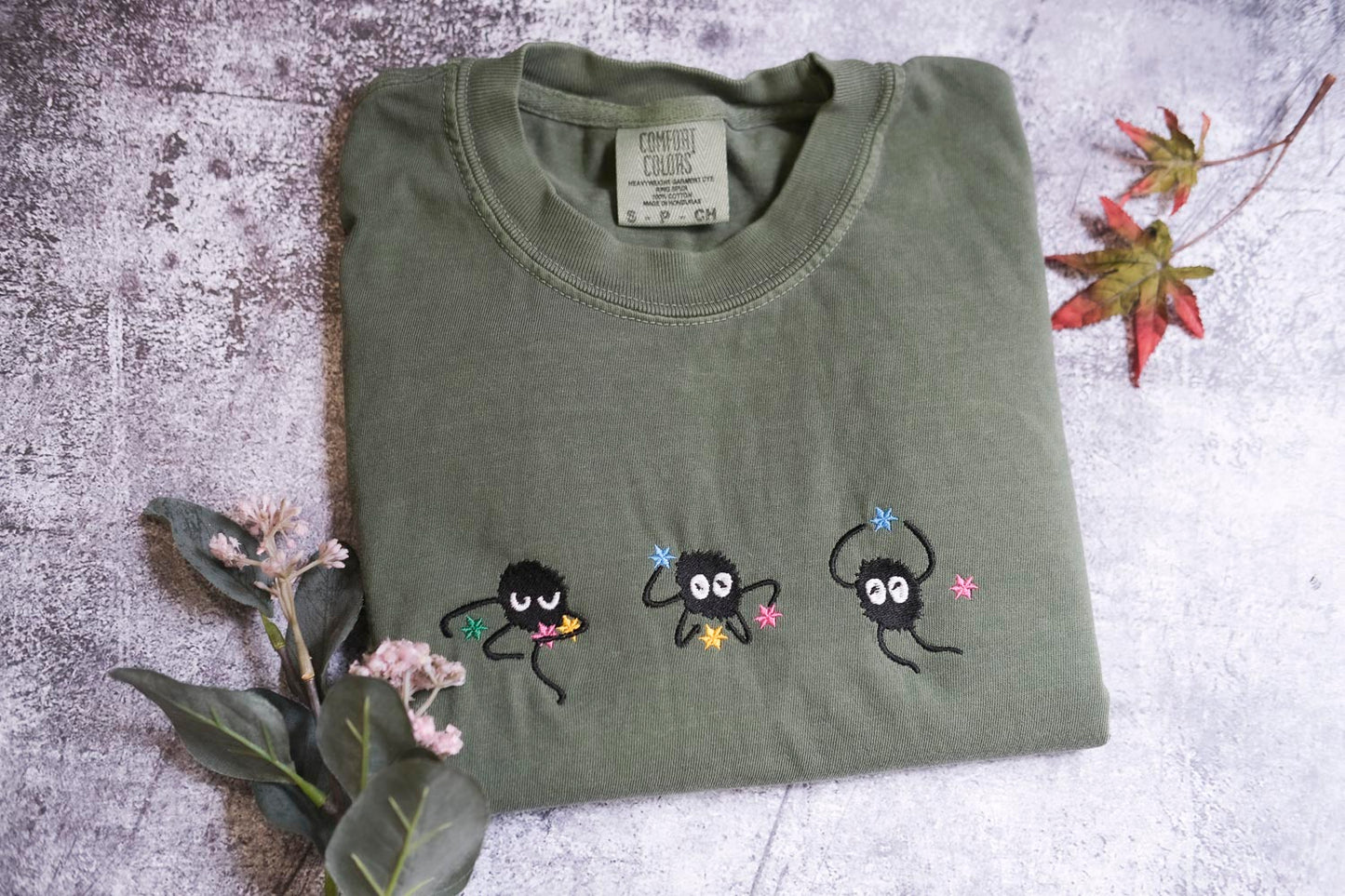 a green t-shirt with three small black creatures with colorful eyes and arms, along with some flowers and leaves on a textured surface.