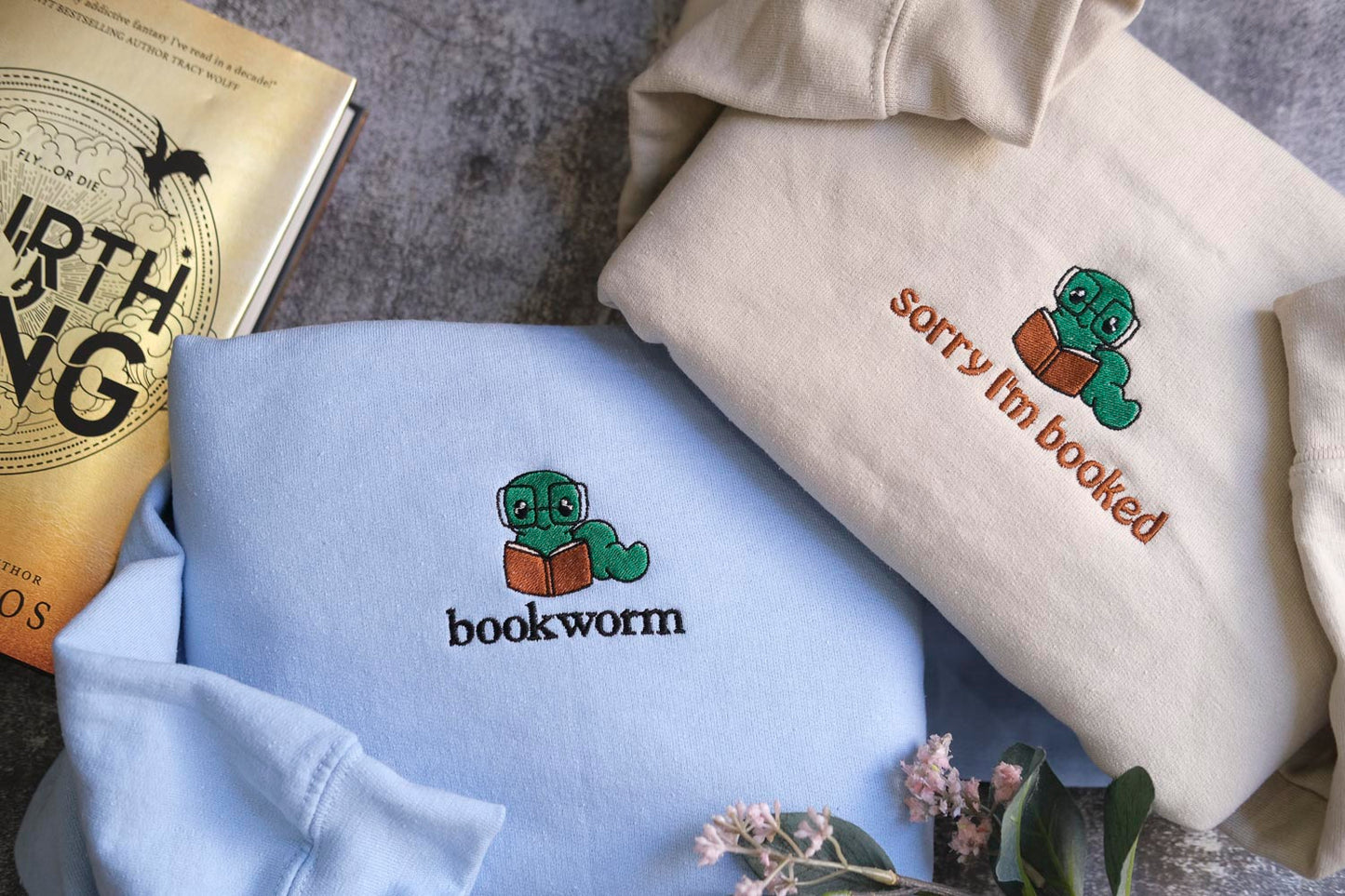 two sweatshirts with a bookworm design, one light blue and one beige, along with a book titled "The North Star" and a book titled "Sorry I'm Booked".