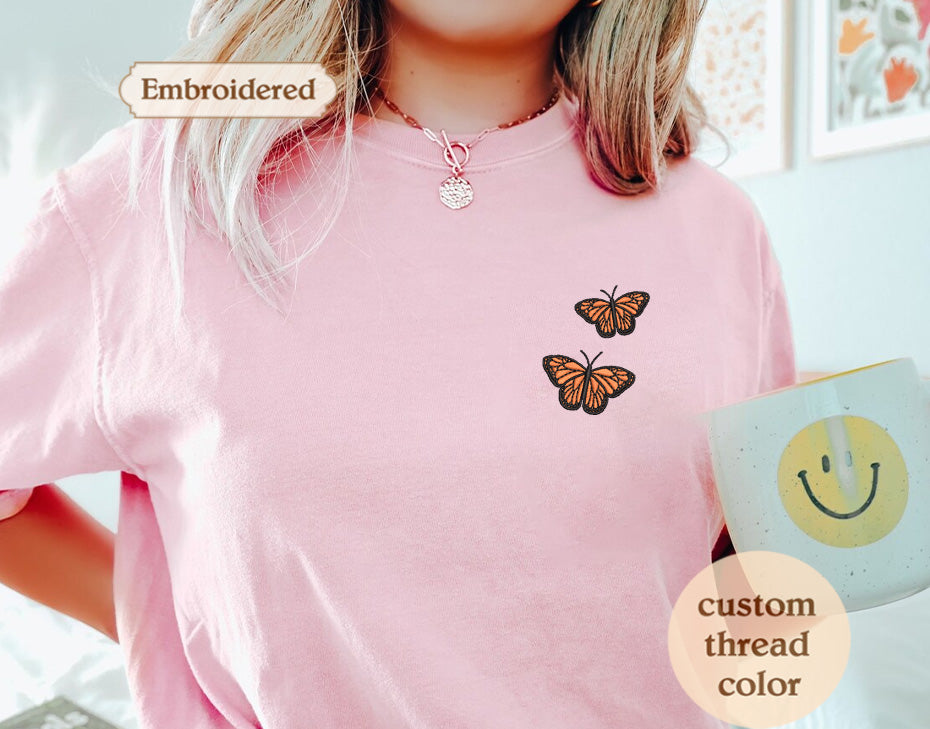 A person wearing a pink t-shirt with a butterfly embroidered on it, holding a mug with a smiley face design.