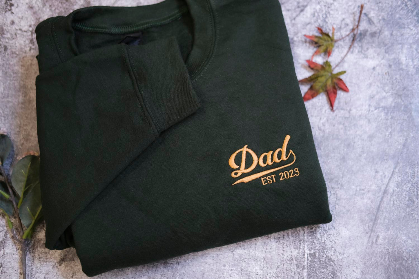 A dark green long-sleeved shirt with a gold text logo that says "DAD EST 2023" on it.