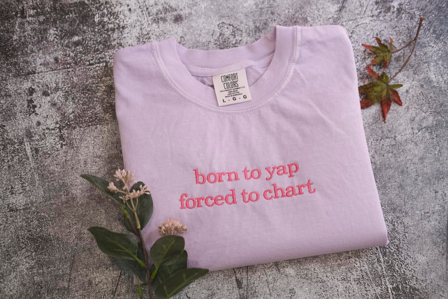 A pink t-shirt with the text "born to yap forced to chart" printed on it, along with some leaves and flowers.