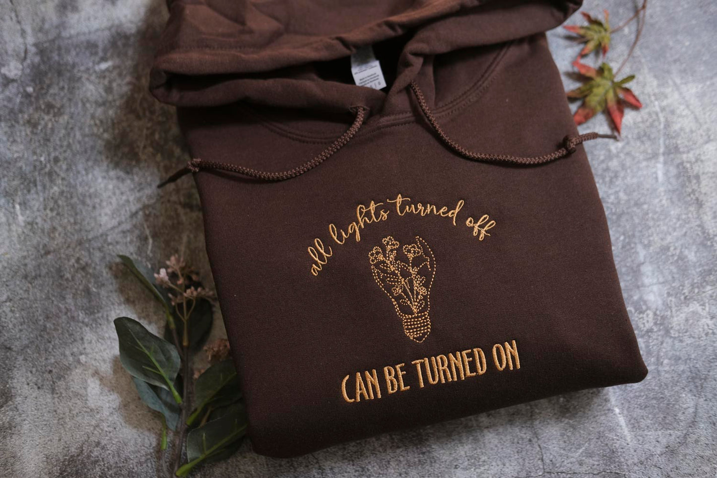 A brown hoodie with a gold embroidered design and text on it, placed on a gray surface with some leaves and a plant.