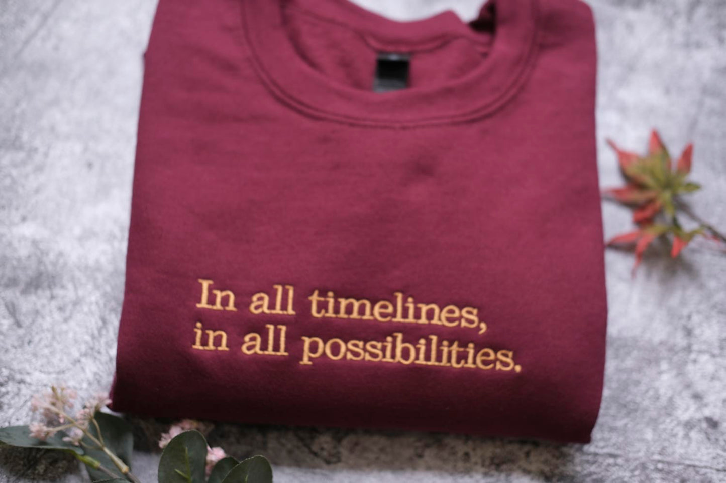 A maroon t-shirt with the text "In all timelines, in all possibilities" printed on it, along with a small leaf and a plant.