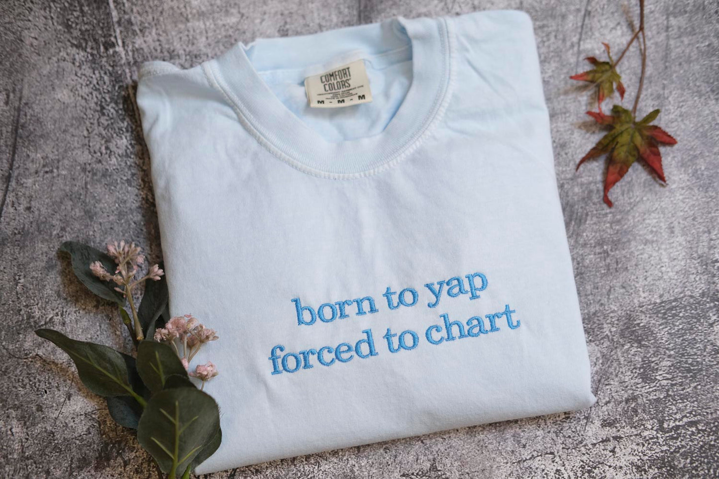 A white t-shirt with the text "born to yap forced to chart" printed on it, along with some leaves and flowers.