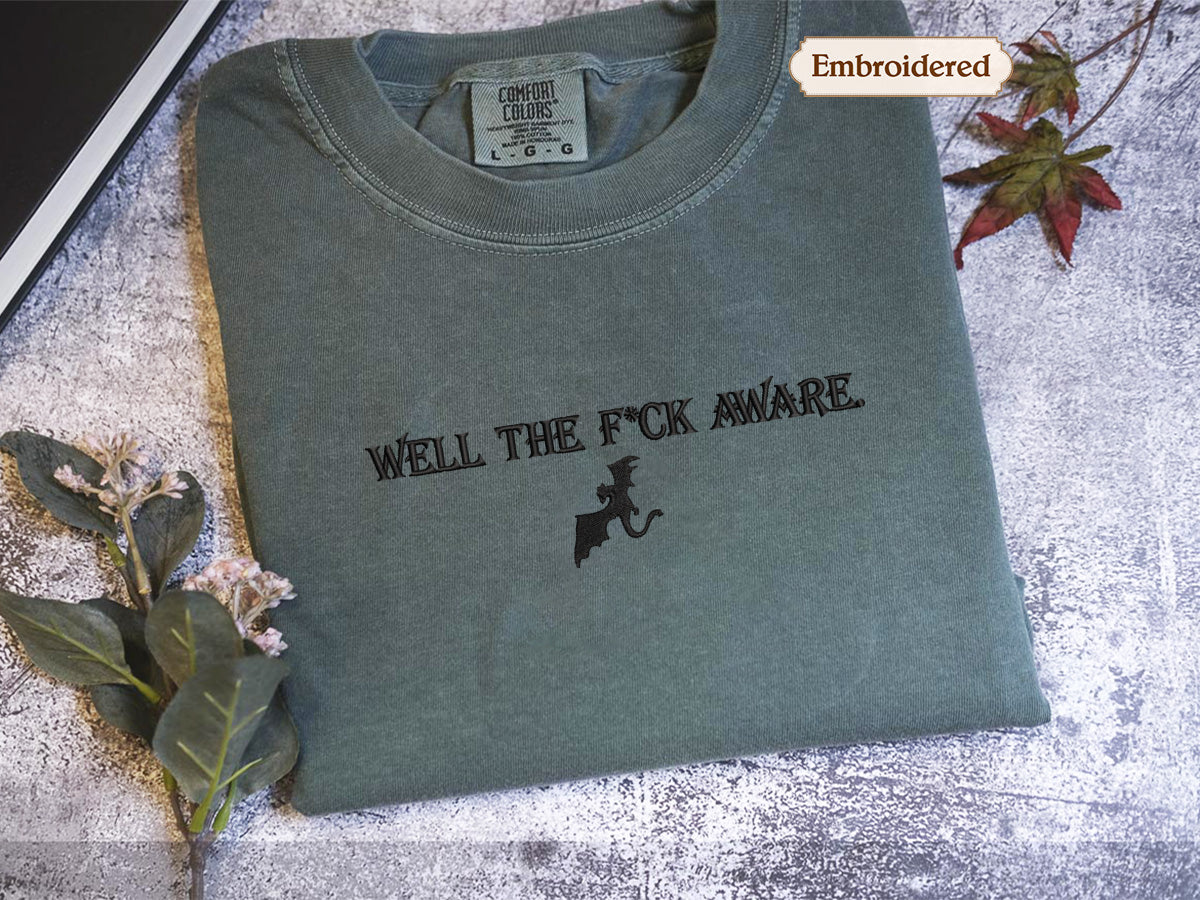 a gray t-shirt with the text "WELL THE F*CK AWARE" printed on it, along with a graphic of a dragon. The t-shirt is placed on a surface with some leaves and flowers, and there is a small embroidered label visible on the collar of the shirt.