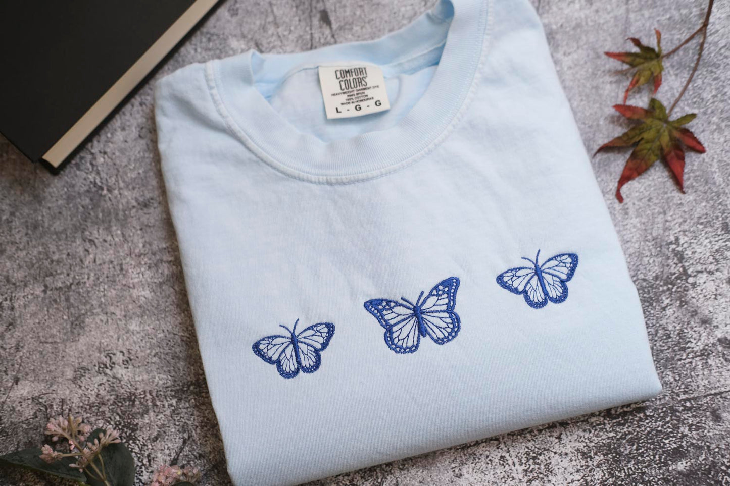 a white t-shirt with three blue butterflies printed on it, placed on a gray surface alongside some leaves and a book.