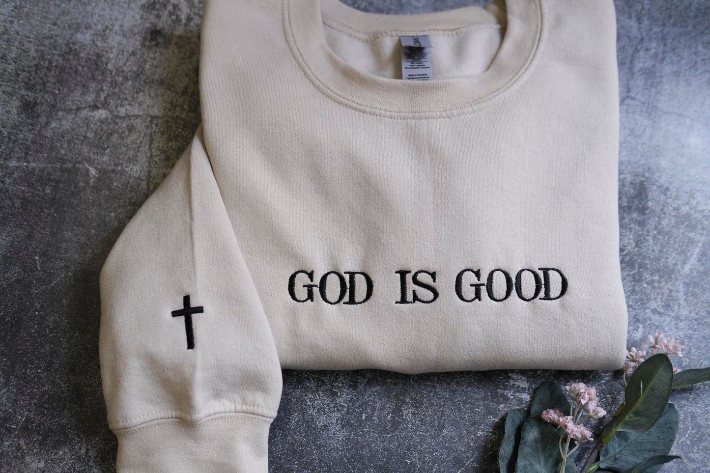 A beige sweatshirt with the text "GOD IS GOOD" printed on it, along with a small black cross symbol on the left sleeve.