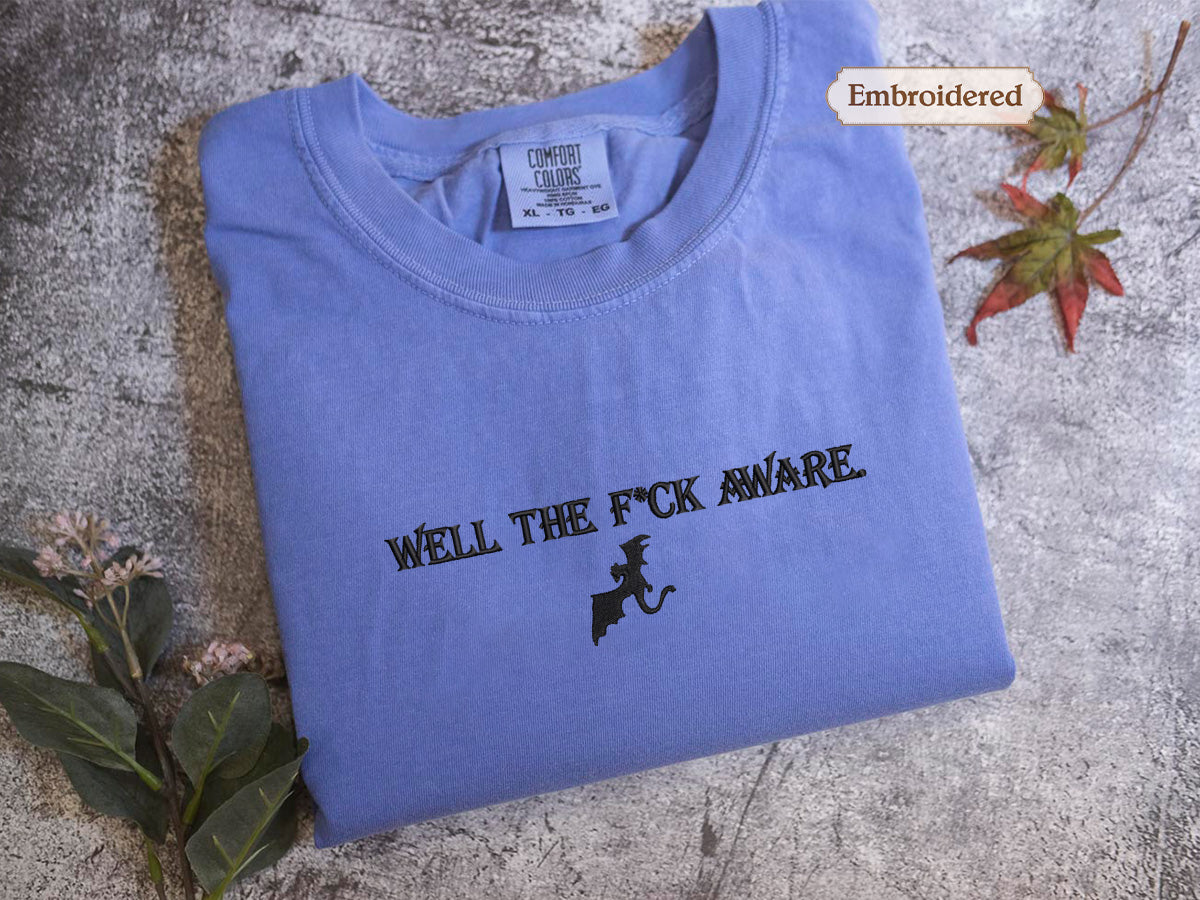 A blue t-shirt with the text "WELL THE F*CK AWARE" printed on it, along with a graphic of a cat. The t-shirt is folded and placed on a surface with some leaves and a small plant.