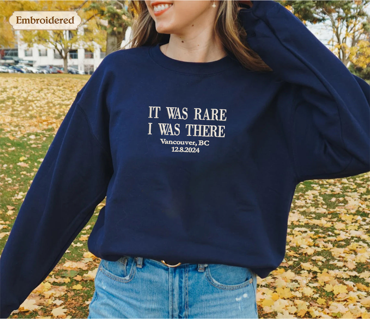 A woman wearing a navy blue sweatshirt with the text "IT WAS RARE I WAS THERE" printed on it, standing in a field with fallen leaves.
