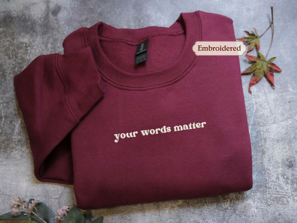 A burgundy sweatshirt with the text "your words matter" embroidered on it, along with a small leaf and a tag.