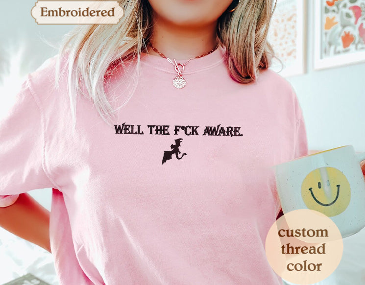 A person wearing a pink t-shirt with the text "WELL THE FICK AWARE" printed on it, along with a small graphic of a dragon. They are holding a mug with a smiley face on it.