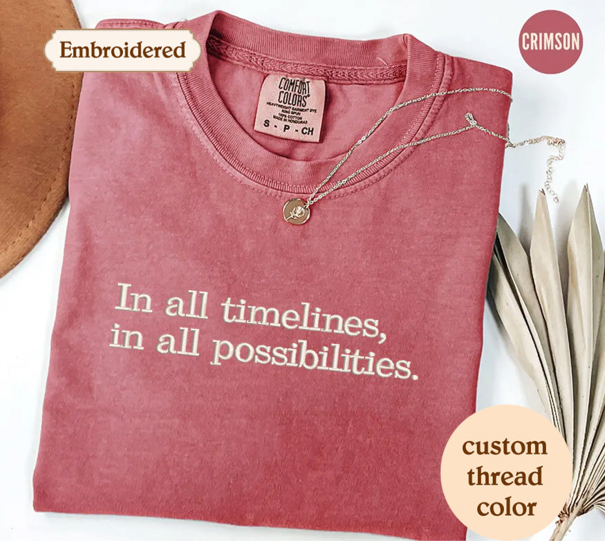a pink t-shirt with the text "In all timelines, in all possibilities" embroidered on it. The t-shirt is displayed on a white background, with a necklace and some leaves visible in the foreground.