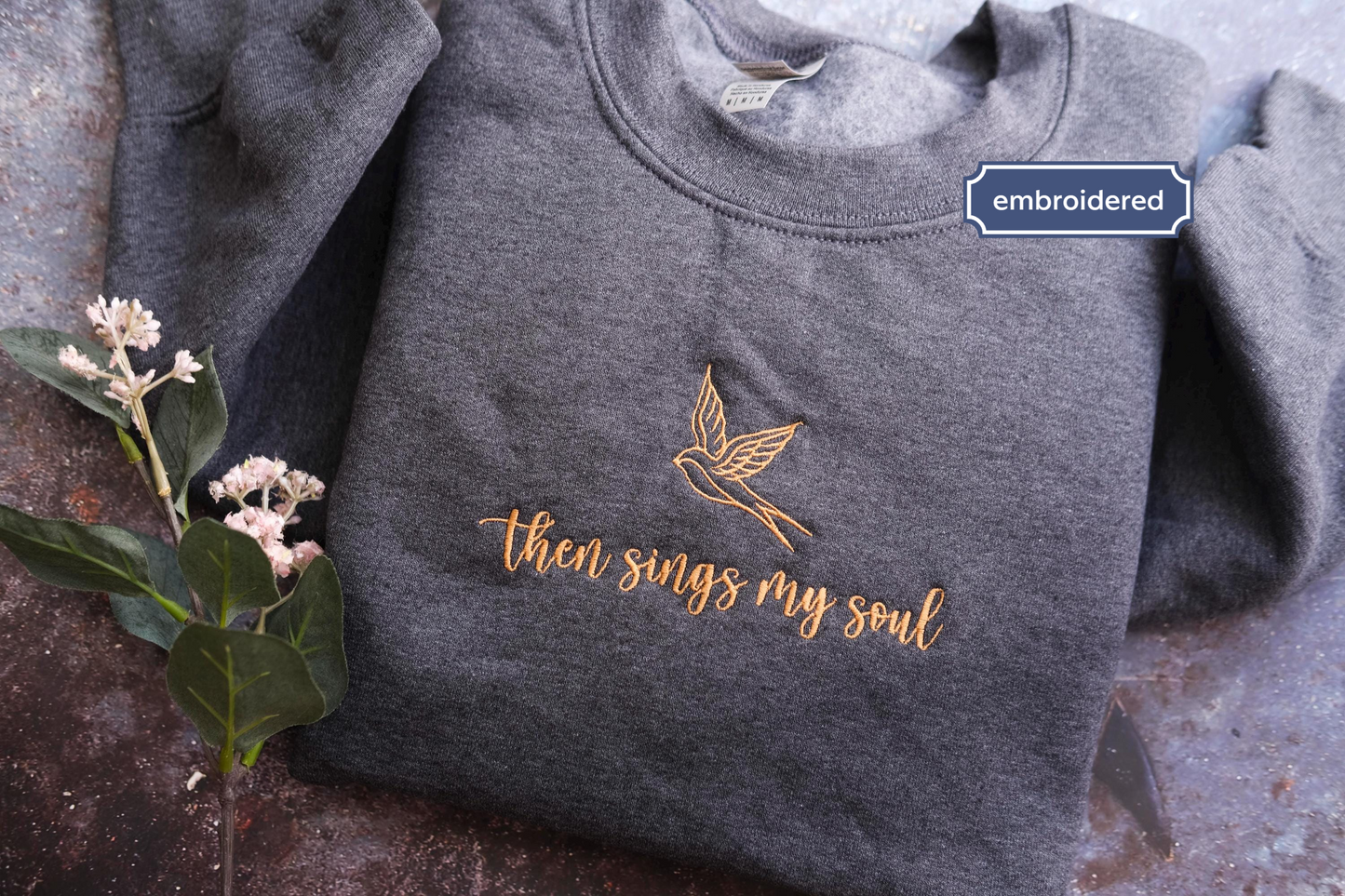 A gray sweatshirt with a gold embroidered design and text, along with a small plant and a blue embroidered label.