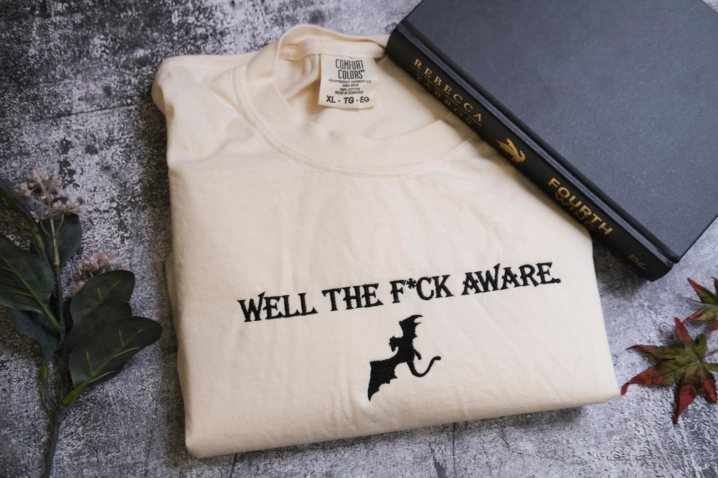 a white t-shirt with the text "WELL THE F*CK AWARE" printed on it, along with a graphic of a bat. The t-shirt is placed next to a book titled "REBECCA" and a plant.