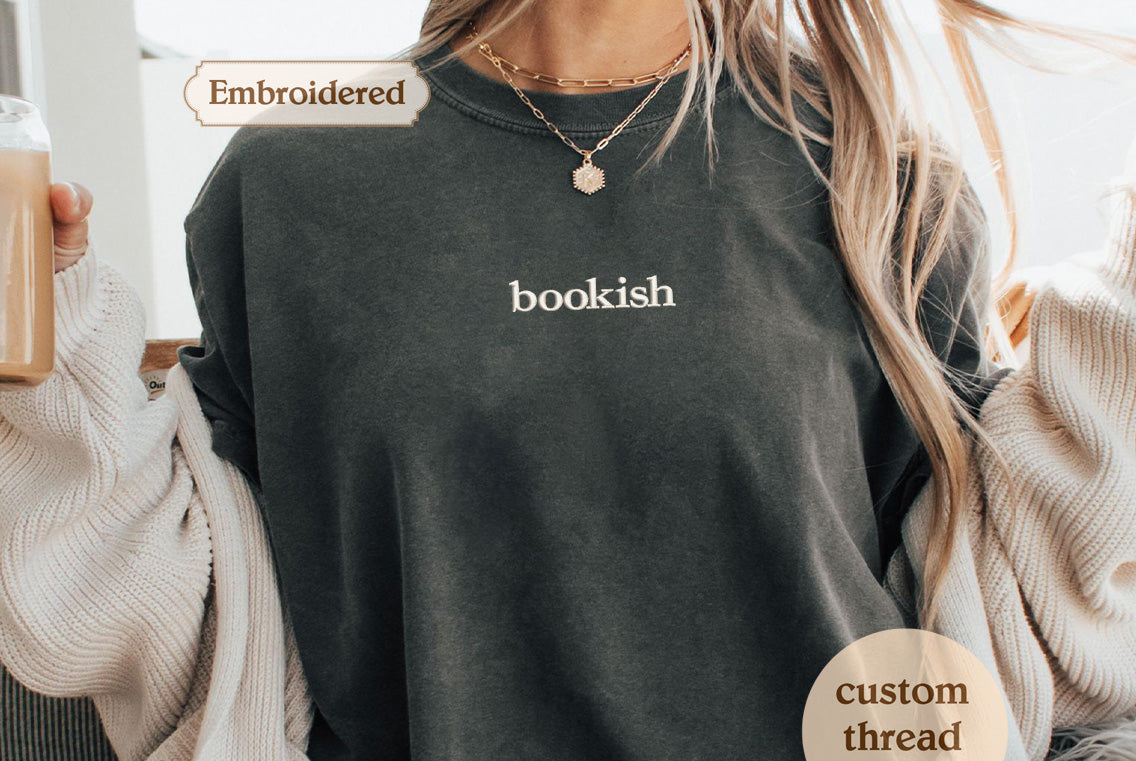 A person wearing a gray t-shirt with the word "bookish" printed on it, along with a necklace and a sweater.