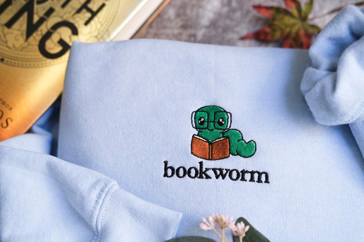 a light blue sweatshirt with a small green book worm wearing glasses and holding a book, along with the word "bookworm" printed on it.
