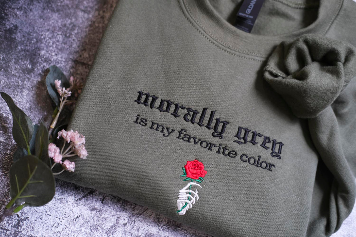 A gray sweatshirt with the text "morally grey is my favorite color" and a red rose embroidered on it, along with a skeleton hand holding the rose.