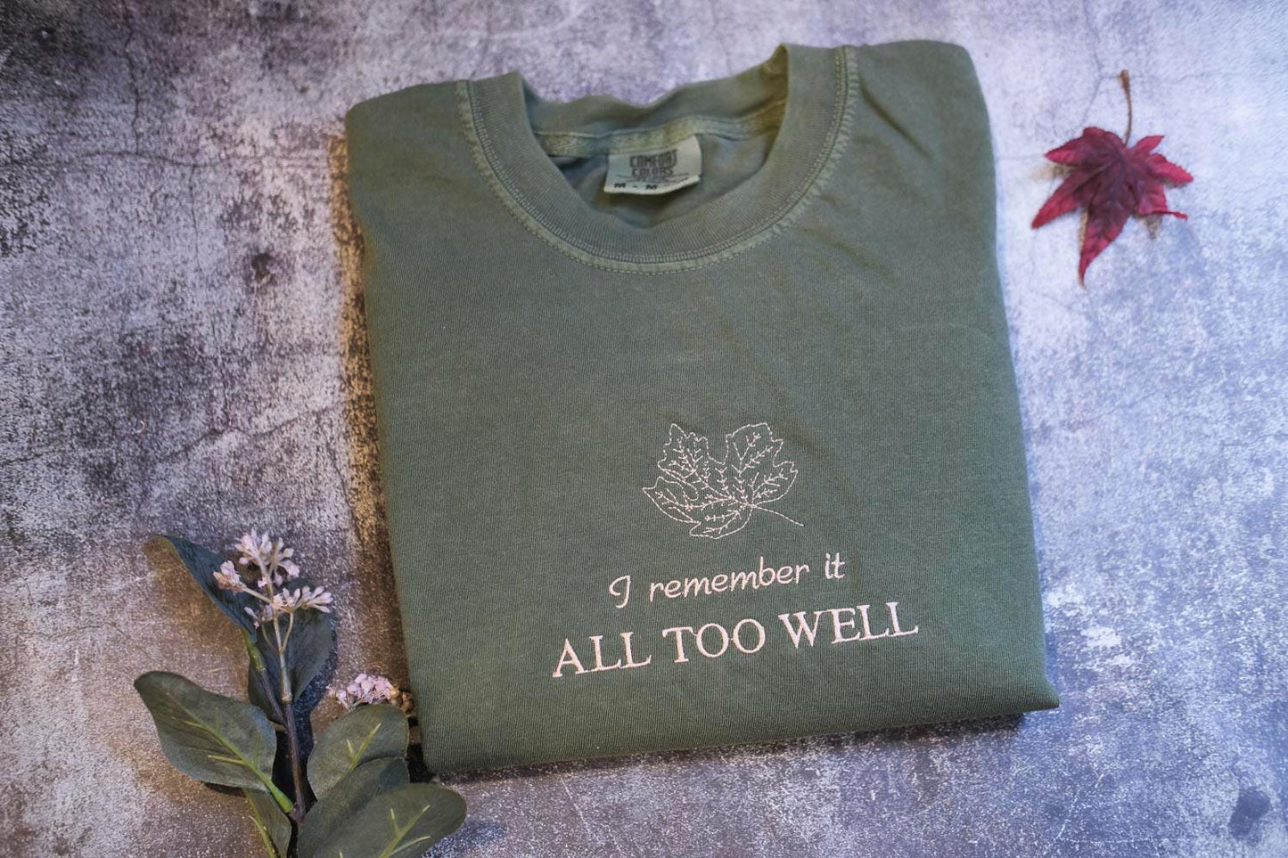 a green t-shirt with the text "I remember it all too well" printed on it, along with a leaf design. The t-shirt is placed on a gray surface, and there is a red leaf and some flowers nearby.